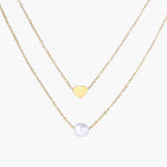 Two delicate gold-plated chains with heart charm and white pearl bead on cream background, minimal layered design.