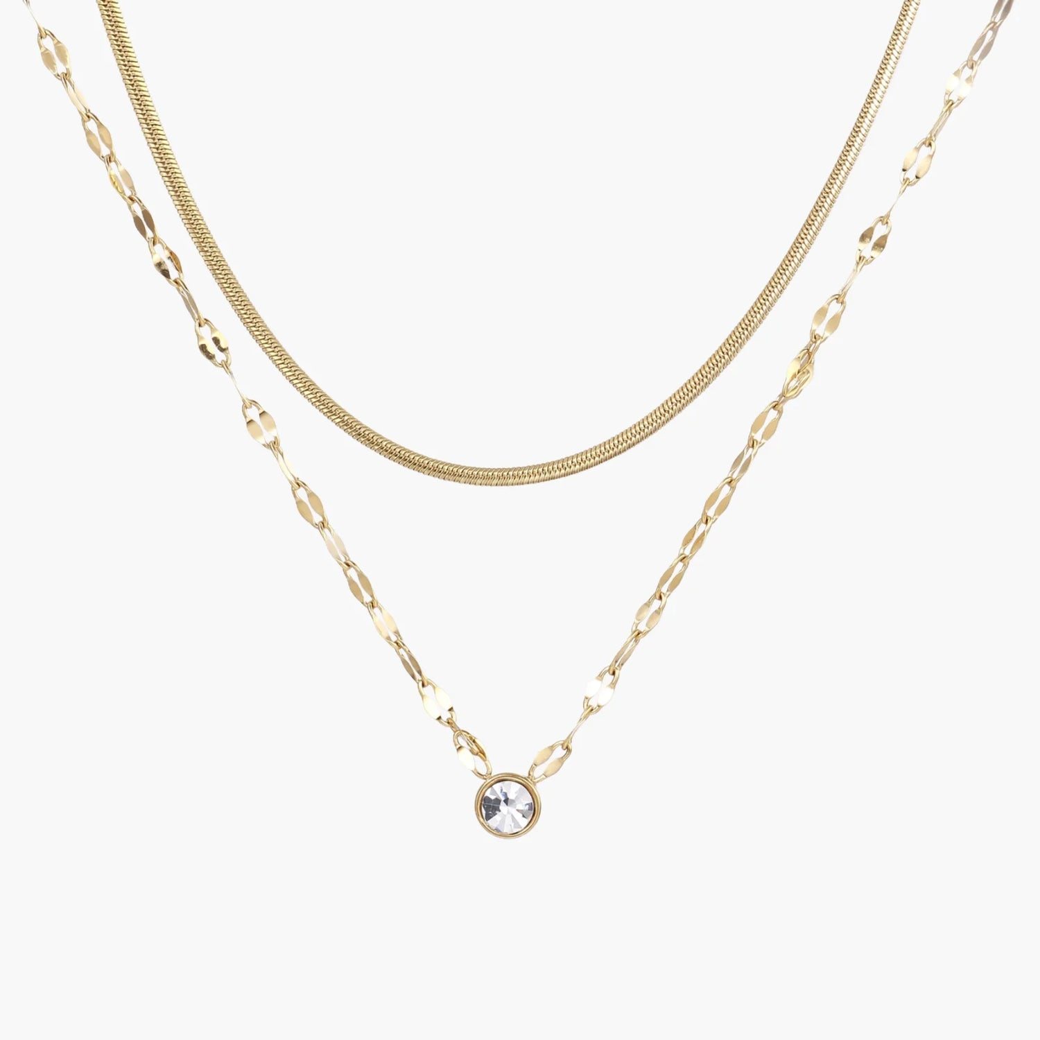Layered gold necklace with herringbone and link chains, small bezel-set crystal pendant on cream background